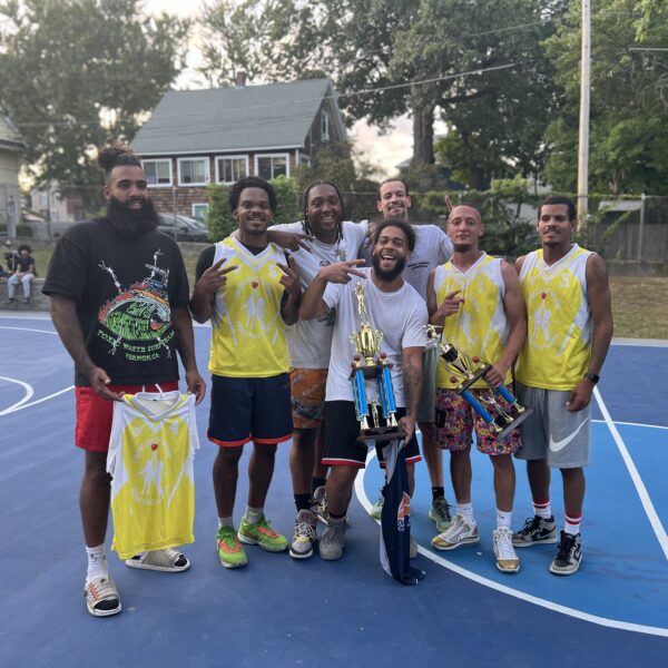 A group of seven men posing on a basketball court with trophies.